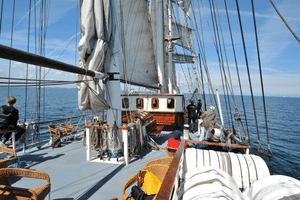 Deck of Mare Frisium with white sails, rigging, wicker chairs, and passengers. Blue sea and sky visible in background.