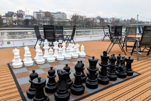 Sonnendeck der MS Classica mit übergroßem Schachspiel auf Holzdeck. Schwarze Klappstühle um das Brett mit Fluss und Stadtpanorama im Hintergrund.