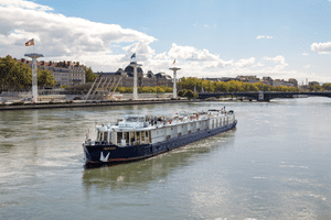 Flusskreuzfahrtschiff Provence auf der Rhône mit blauem Rumpf und weißem Aufbau. Französische Flaggen sichtbar, Stadtufer mit Gebäuden im Hintergrund.