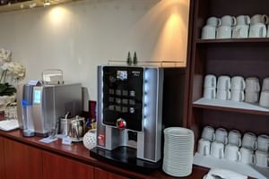 Kaffeestation mit automatischer Espressomaschine und Wasserspender auf Holztheke. Weiße Tassen und Teller auf Regalen darüber.