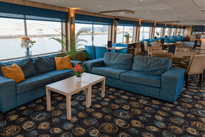 Spacious lounge on DE Nassau ship with teal sofas, patterned carpet, and large windows overlooking the water. Modern interior with plants and flowers.