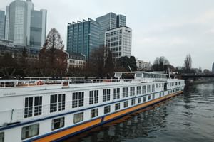 Weißes Flusskreuzfahrtschiff MS Classica mit blauem und orangem Zierstreifen am Fluss vertäut, mit modernen Glasgebäuden und kahlen Bäumen im Hintergrund.