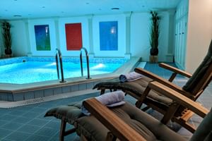 Innenpool mit blauen Mosaikfliesen und Holzliegen mit Handtüchern. Drei farbige Wasserspiele an der Wand und Dekorationspflanzen.