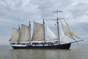 Four-masted sailing ship Leafde fan Fryslân with dark hull and cream-colored sails on calm water under cloudy sky.