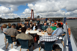 Group of passengers seated at tables on the sun deck of ship De Nassau. Red bridge and river visible in background under cloudy sky.
