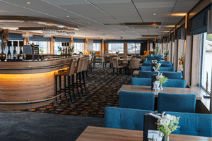 Modern lounge on MS Nassauer with curved wooden bar, blue upholstered seating, and large windows overlooking the water.