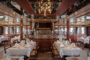 Elegantes Schiffsrestaurant mit weißen Tischdecken, Holzstühlen und historischem Schaufelrad als Mittelpunkt. Spiegeldecke und warme Beleuchtung.
