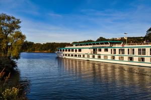 MS Switzerland river cruise ship with white hull and green-turquoise trim sailing on calm river under blue sky, surrounded by trees.
