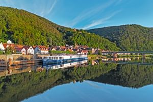 River cruise ship Casanova moored along a riverside village with colorful houses, forested hills, and perfect water reflections under blue sky.