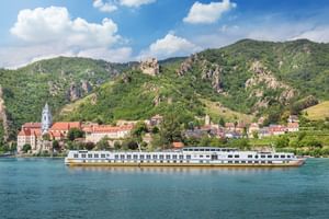 Weißes Flusskreuzfahrtschiff MS Vista Classica auf der Donau vor historischer Stadt mit Kirchtürmen und grünen Bergen mit Burgruine.