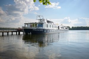 White river cruise ship MS Princess moored at wooden dock on calm water under blue sky with white clouds and green trees in background.
