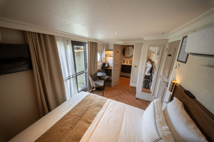 Elegant river cruise suite with double bed, balcony door, seating area, wardrobe, and warm wood flooring on the Magnifique X upper deck.