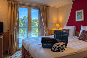 Double cabin on Casanova upper deck with two beds, balcony door, river view, beige walls, burgundy accent wall, and cycling equipment on bed.