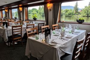 Eleganter Speisesaal an Bord der Classica mit weiß gedeckten Tischen, Holzstühlen und großen Fenstern mit Blick auf Fluss und grüne Landschaft.