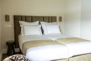 Modern hotel bedroom with double bed, white linens, beige runner, padded headboard, and bronze wall sconces in Magnifique X Junior Suite.