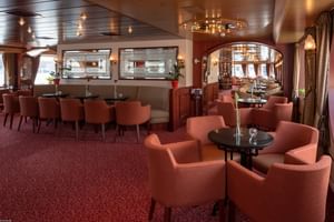 Eleganter Salon an Bord des Schiffes Switzerland mit korallfarbenen Sesseln, runden Tischen, rotem Teppich und holzgetäfelter Decke mit Kronleuchtern.