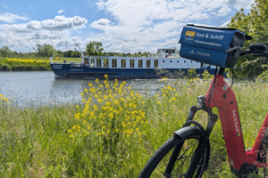 Flusskreuzfahrtschiff Casanova mit blau-weißem Rumpf auf Wasserstraße. Rotes Fahrrad mit Reiseveranstalter-Schild im Vordergrund, gelbe Wildblumen am Ufer.