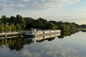 White Princess river cruise ship moored at a wooden dock in Schwedt. The vessel is reflected in the calm water, surrounded by green trees under a cloudy sky.