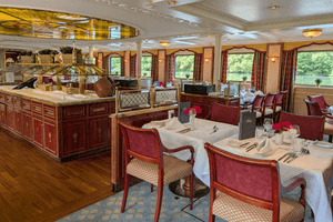 Elegant restaurant aboard Casanova with burgundy upholstered chairs, white-clothed tables, wooden buffet counter, and large windows overlooking water.