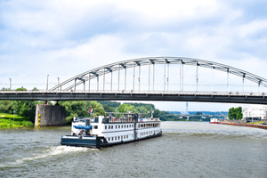 White paddle steamer De Nassau sailing on a river, passing under a steel arch bridge. Green riverbanks and cloudy sky visible.
