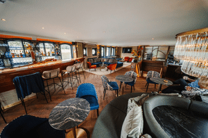 Spacious lounge on Magnifique X with wooden bar, blue velvet chairs, patterned seating, red armchairs, and elegant chandelier. Large windows line the walls.