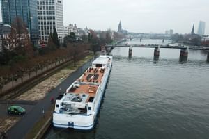 Weißes Flusskreuzfahrtschiff MS Classica am städtischen Flussufer mit Brücken und Stadtsilhouette im Hintergrund unter bewölktem Himmel.