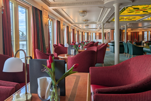 Eleganter Salon auf dem Schiff Casanova mit rot gepolsterten Stühlen, runden Tischen mit Tulpen, großen Fenstern, weißen Säulen und Zierdecke.