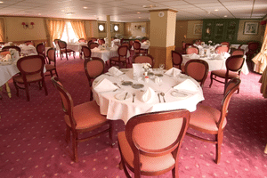 Eleganter Speisesaal auf dem Schiff Provence mit runden Tischen mit weißen Tischdecken, burgunderroten Polsterstühlen und rosa gemustertem Teppich.