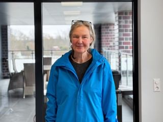 Monika Huber wearing a bright blue jacket and sunglasses on her head, smiling in an indoor setting with glass doors and terrace visible behind.