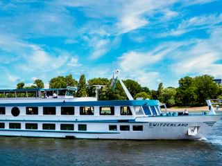 White river cruise ship MS Switzerland sailing on calm water with blue sky, trees and greenery visible along the shore in the background.