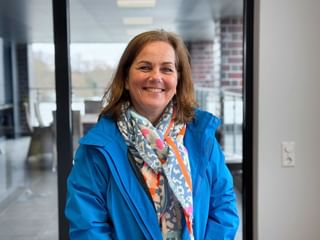 Michaela Kuhn smiling, wearing a bright blue jacket and colorful floral scarf, standing in a modern office with glass walls.