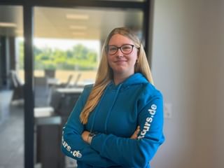 Lea Sophie Döscher mit langen blonden Haaren und Brille, trägt einen blauen Hoodie mit weißem Text auf den Ärmeln, steht in einem modernen Büro.