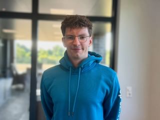 Jannik Rauchfleisch mit Brille und blauem Hoodie, stehend in einem modernen Büro mit Glastüren und Fenstern im Hintergrund.