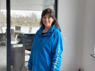 Katrin Volkmann with dark hair wearing a bright blue jacket, smiling in a modern interior with large windows showing outdoor scenery.