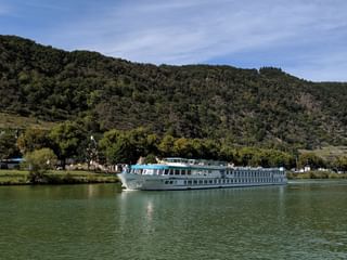 Weißes Flusskreuzfahrtschiff Switzerland auf ruhigem grünen Wasser mit bewaldeten Hügeln und kleinem Dorf im Hintergrund unter blauem Himmel.