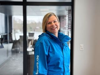 Maxi Loska wearing a bright blue jacket, smiling warmly in an indoor setting with glass doors and modern office space visible in the background.