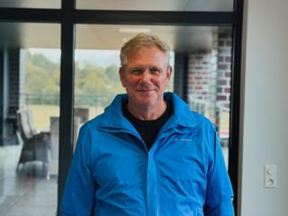 Karsten Frischknecht lächelnd in blauer Outdoorjacke und schwarzem Pullover, stehend in Innenräumen mit Fenstern und Landschaft im Hintergrund.
