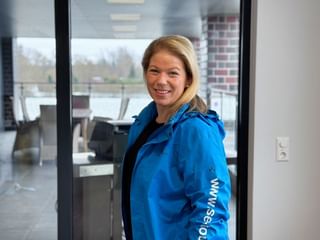 Victoria Vari wearing a bright blue jacket with white text on the sleeve, smiling at the camera in a modern office setting with windows.