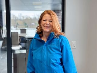 Ulrike Elster with long reddish-blonde hair wearing a bright blue jacket, smiling in a modern office with glass walls and windows.