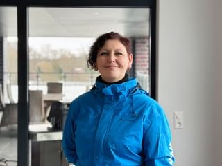 Monika Fürdos wearing a bright blue outdoor jacket, smiling in an indoor setting with large windows showing an outdoor view.