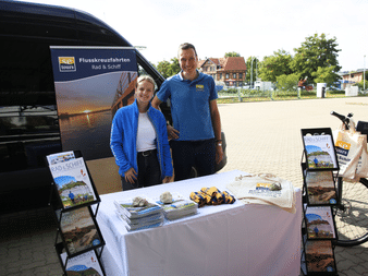 Zwei Mitarbeiter in blauen Shirts an einem Außenstand mit Broschüren und Werbematerialien für Flusskreuzfahrten auf einem weiß gedeckten Tisch.