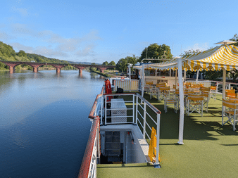 Sun deck of river cruise ship Casanova with yellow-white striped awning, wooden chairs, and green artificial turf. River and bridge visible in background.