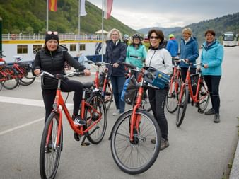Gruppe von Radfahrern mit roten Leihrädern an einem Flusshafen an der Donau. Weißes Passagierschiff und bewaldete Hügel im Hintergrund.