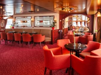 Elegant salon bar on MS Switzerland with red carpeting, orange upholstered chairs, round tables, and decorative mirrors on walls.