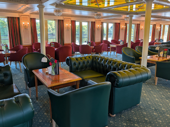 Eleganter Salon an Bord der Casanova mit grünen Chesterfield-Ledersofas, roten Samtstühlen, Holztischen, weißen Säulen und großen Fenstern.