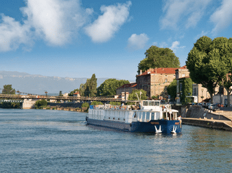 Weiß-blaues Flusskreuzfahrtschiff MS Provence auf der Rhône mit Brücke und historischen Gebäuden umgeben von Bäumen im Hintergrund.