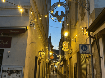 Schmale Kopfsteinpflastergasse Drosselgasse in Rüdesheim mit Lichterketten, traditionellen Schildern und Kirchturm im Hintergrund in der Dämmerung.