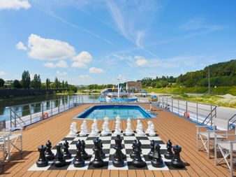 Sun deck of MS SE-MANON featuring a giant chess set on checkered floor, swimming pool, wooden deck, and river view with green hills.