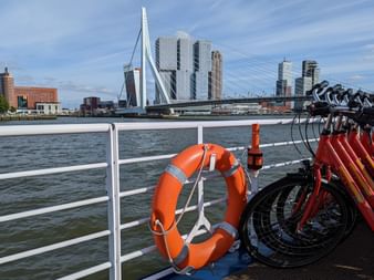 Blick vom Schiffsdeck in Rotterdam mit orangem Rettungsring, roten Fahrrädern und Skyline mit Erasmusbrücke und modernen Wolkenkratzern.