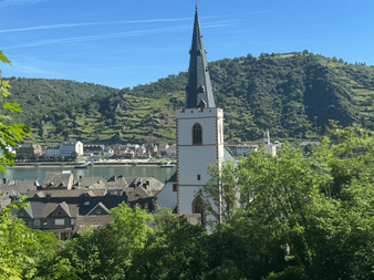 Weißer Kirchturm mit Spitze in St. Goar mit Blick auf den Rhein. Weinberge bedeckte Hügel und Stadtgebäude im Hintergrund sichtbar.